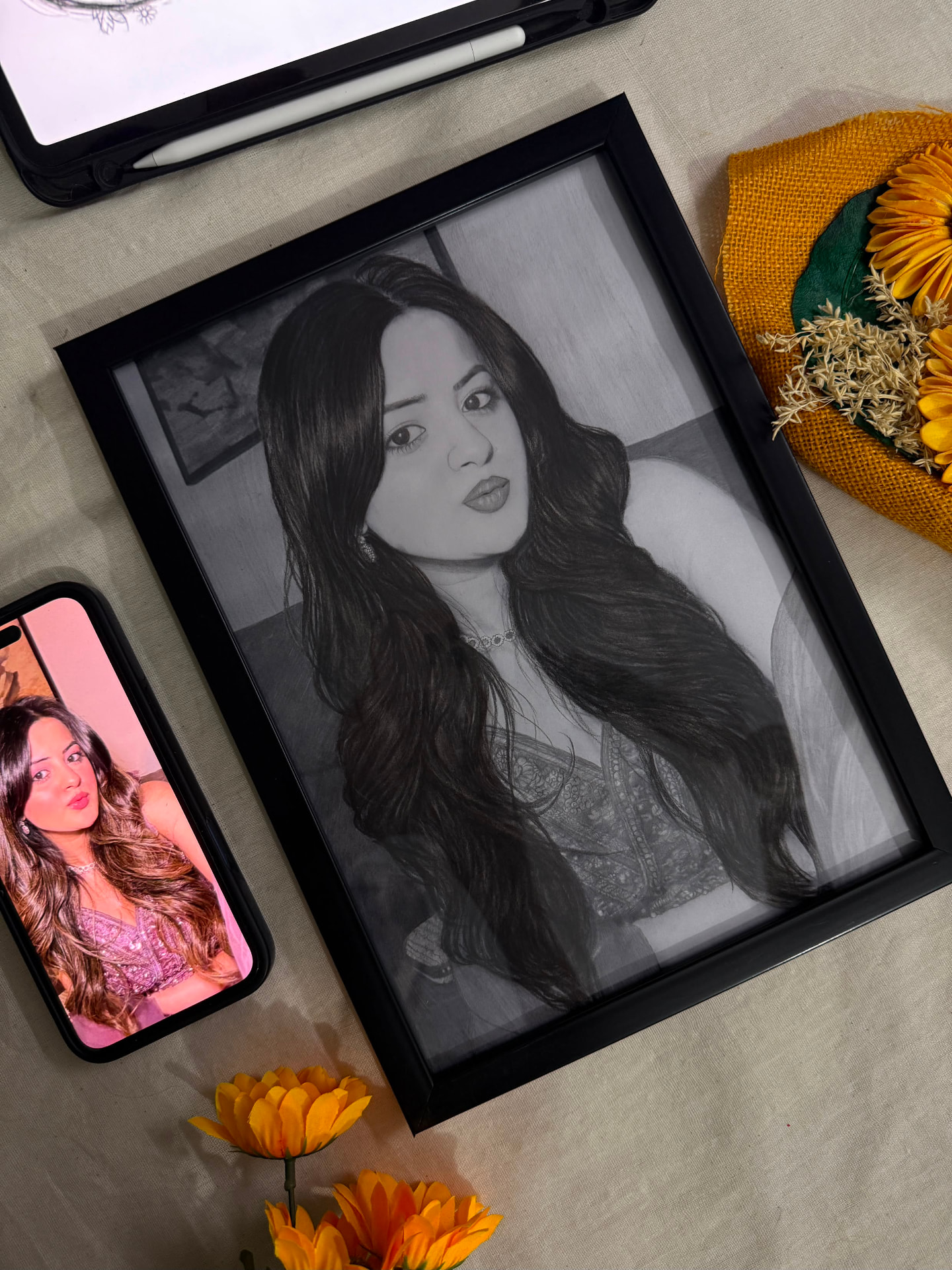 Framed graphite portrait displayed beside its reference photo and flowers